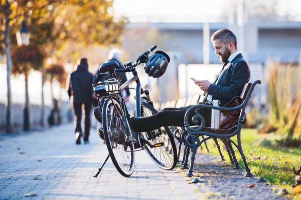 Empresario viajero con bicicleta sentado en el banco de la ciudad, utilizando el teléfono inteligente . — Foto de Stock