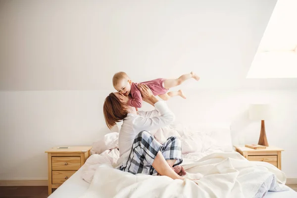 Une jeune mère avec une petite fille assise à l'intérieur sur le lit le matin, jouant . — Photo