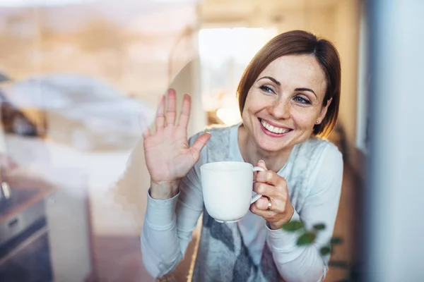 A young woman with cup of coffee looking out of a window, waving goodbye. — Stock Photo, Image