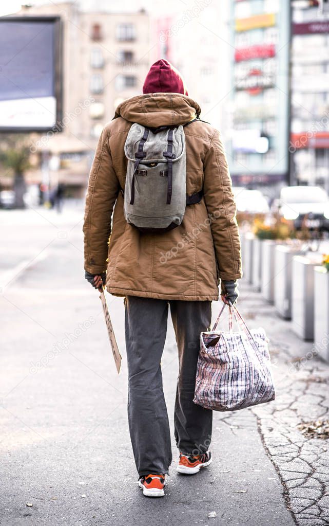 A rear view of homeless beggar man walking outdoors in city, holding bag.