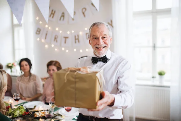 En äldre man håller en present i en ruta på en inomhus familj födelsedagsfest. — Stockfoto