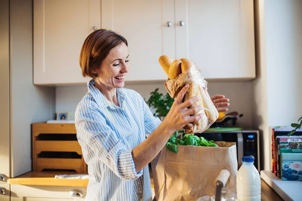 A young woman standing indoors in kitchen, unpacking shopping bag. — Stock Photo, Image