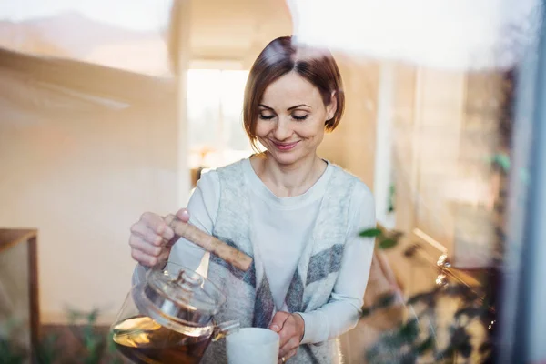 A young woman standing indoors in kitchen, pouring tea. Shot through glass. — Stock Photo, Image