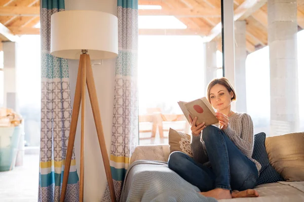 En ung kvinna sitter inomhus på en soffa hemma, läsa en bok. — Stockfoto