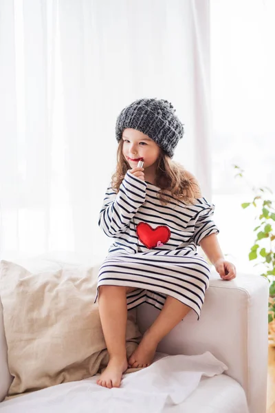 Una pequeña chica feliz en camiseta a rayas en casa aplicando un lápiz labial . — Foto de Stock