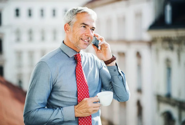 Uomo d'affari maturo con smartphone in piedi su una terrazza in città, facendo una telefonata . — Foto Stock