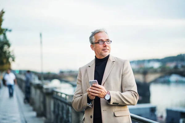 Rijpe zakenman met smartphone permanent door rivier in Praag stad, met behulp van smartphone. — Stockfoto