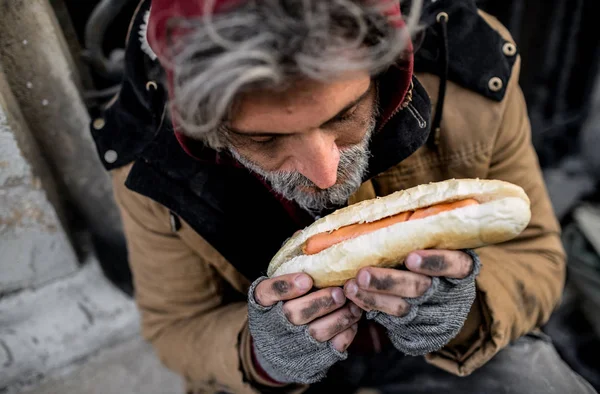 Een bovenaanzicht van dakloze bedelaar man buitenshuis in stad, bedrijf hot-dog. — Stockfoto