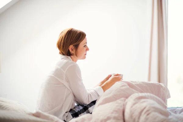 Młoda kobieta z nocy koszuli siedzi wewnątrz na łóżko rano, trzymając kubek. — Zdjęcie stockowe