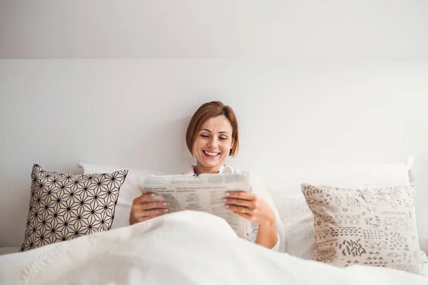 Una joven leyendo periódicos en la cama por la mañana en un dormitorio . — Foto de Stock