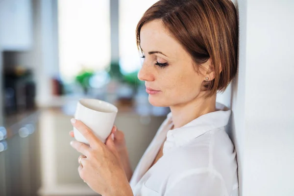 A young woman with cup of coffee standing indoors, leaning on a wall. — Stock Photo, Image
