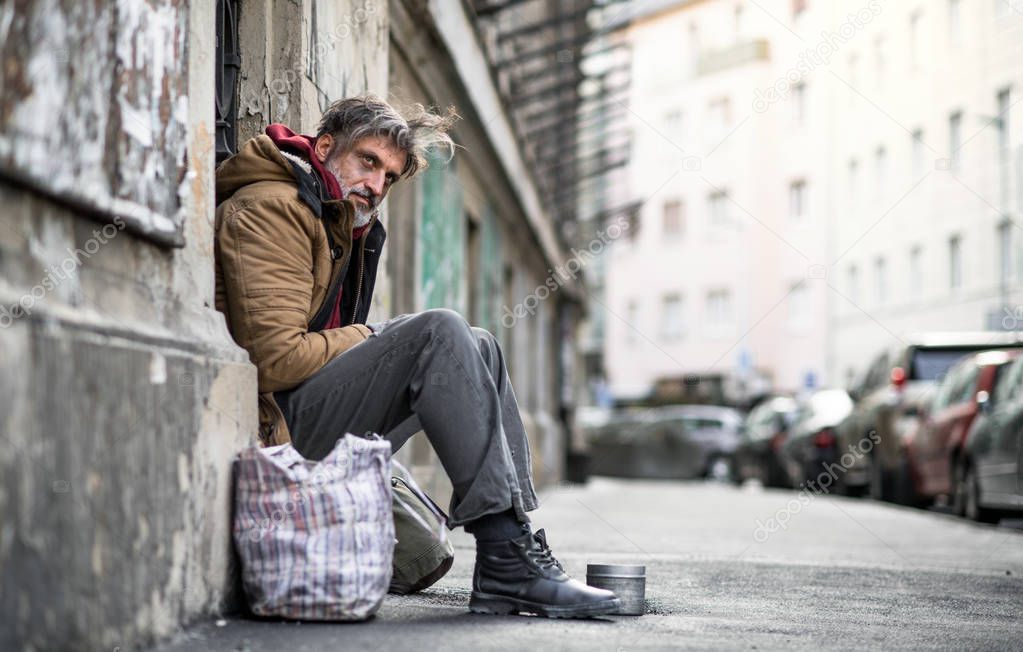 Homeless beggar man sitting outdoors in city, asking for money donation.