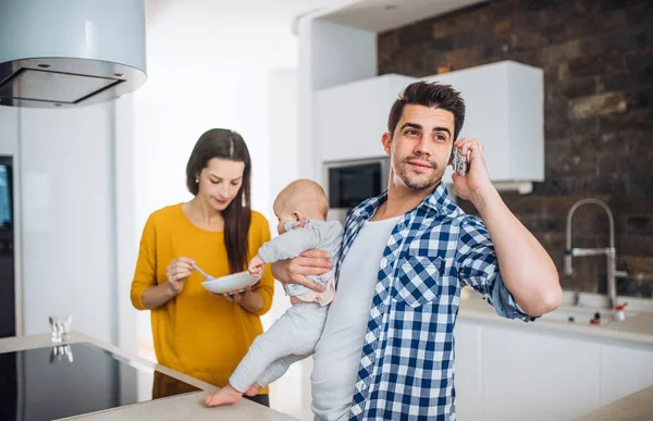 Uma jovem família em casa, um homem fazendo um telefonema e uma mulher alimentando um bebê . — Fotografia de Stock