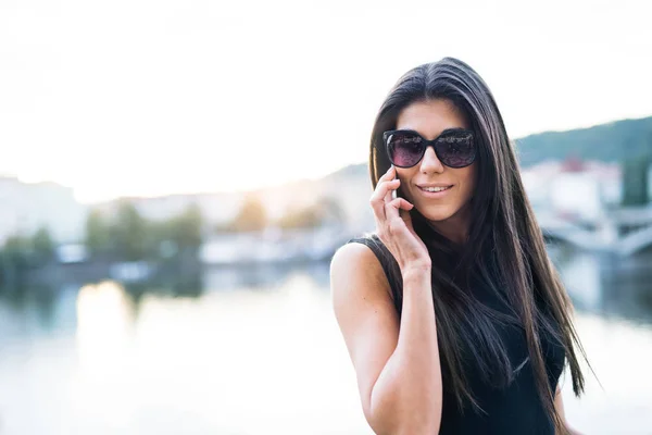 Mulher bonita em vestido preto em pé junto a um rio na cidade de Praga, fazendo um telefonema . — Fotografia de Stock