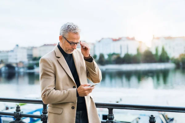 Зрелый бизнесмен со смартфоном, стоящим у реки в Праге, используя смартфон . — стоковое фото