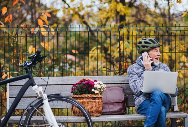 Старший человек с электровелосипедом сидит на скамейке в городе, используя ноутбук и смартфон . — стоковое фото