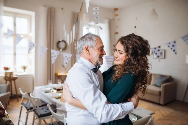 Starší muž a mladá žena stojící uvnitř v místnosti nastavit pro stranu, objímání. — Stock fotografie