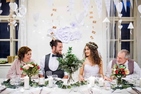 Een jong koppel met ouders zittend aan een tafel op een bruiloft, kijken naar elkaar. — Stockfoto