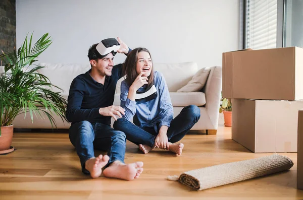 Un jeune couple avec des lunettes VR assis sur le sol, emménageant dans une nouvelle maison . — Photo