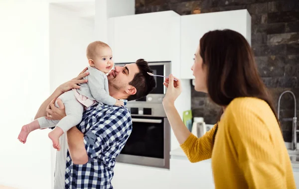 Una giovane famiglia a casa, un uomo che tiene in braccio un bambino e una donna che le dà da mangiare . — Foto Stock