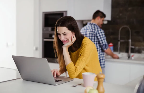 Młoda kobieta korzysta z laptopa w kuchni i mycia naczyń człowiek. — Zdjęcie stockowe