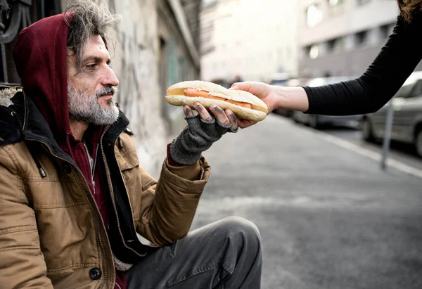 Mulher irreconhecível dando comida para mendigo sem-teto sentado na cidade . — Fotografia de Stock