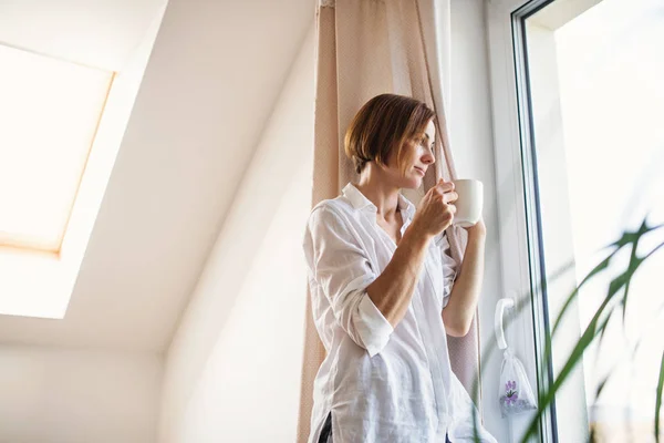 Молодая женщина в ночной рубашке стоит у окна утром, держа чашку кофе . — стоковое фото