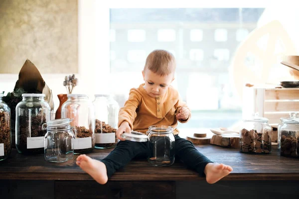 Un pequeño niño descalzo sentado en una mesa en un taller de basura cero . — Foto de Stock