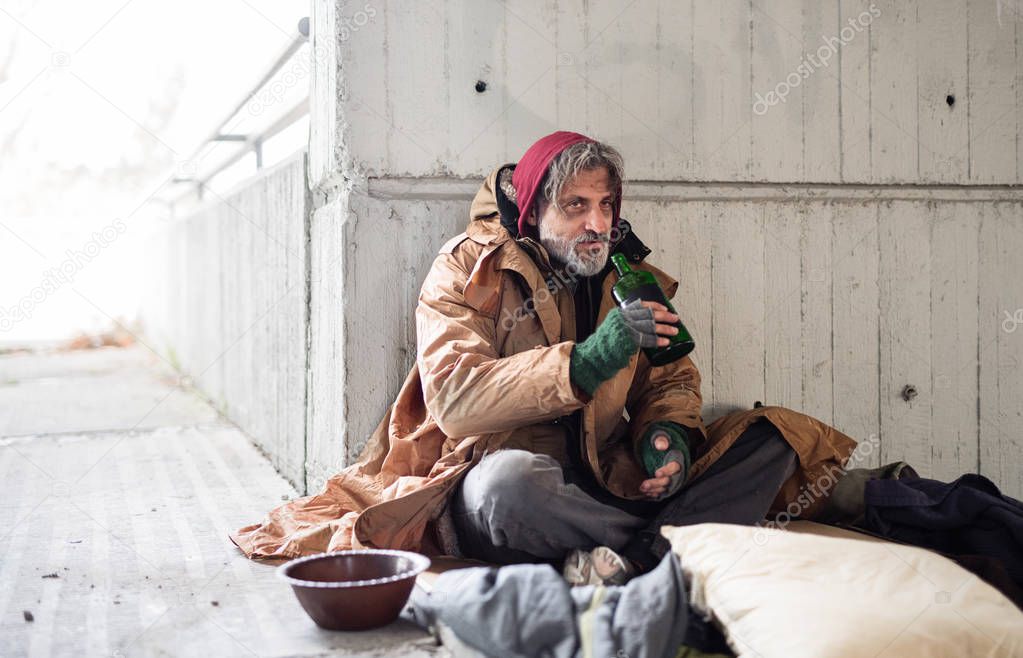 A front view of homeless beggar man sitting outdoors, holding bottle of alcohol. Copy space.