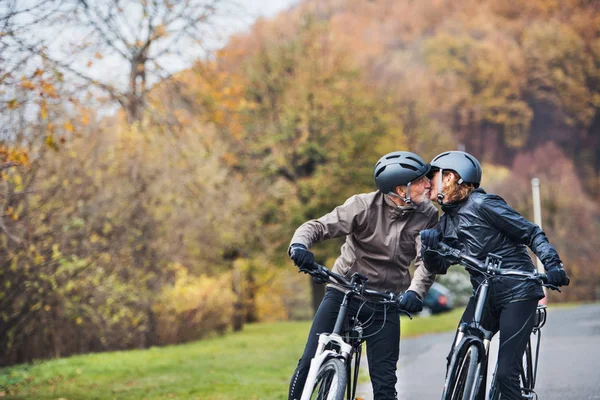 Aktivní senior pár s electrobikes stojící venku na silnici v přírodě, Polibky. — Stock fotografie