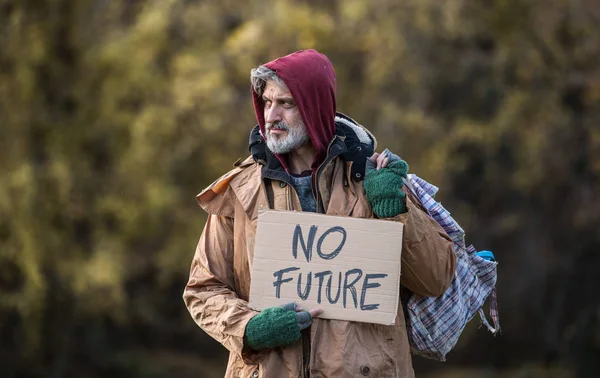 Żebrak bezdomny mężczyzna stojący na zewnątrz, w parku, trzymając worek i karton znak. — Zdjęcie stockowe