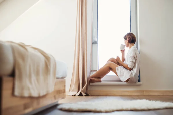 Una joven con camisa de noche sentada junto a la ventana por la mañana . — Foto de Stock