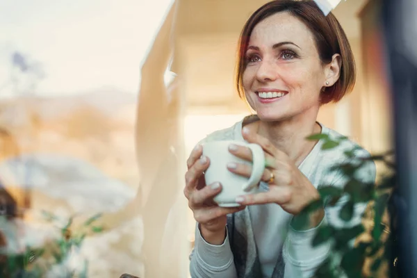 Een jonge vrouw met kopje koffie kijken uit het raam. Shot door glas. — Stockfoto