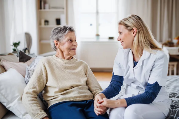 En hälsa besökare att prata med en sjuk äldre kvinna som sitter på sängen hemma. — Stockfoto
