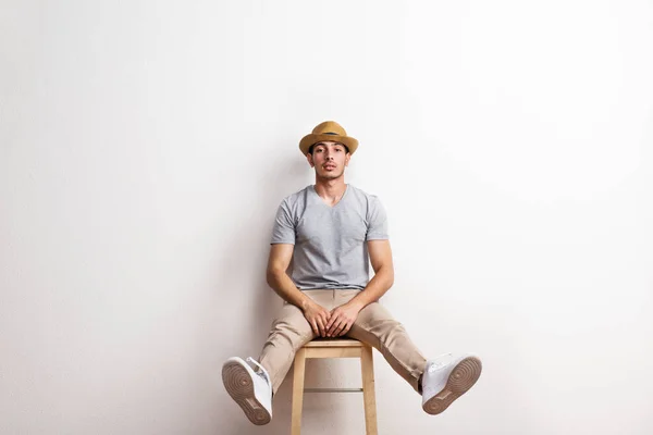 Un joven hispano confiado con sombrero sentado en un taburete en un estudio . — Foto de Stock
