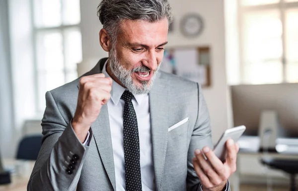 Uomo d'affari maturo con smartphone in piedi in un ufficio, esprimendo eccitazione . — Foto Stock