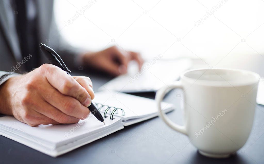 A midsection of businessman sitting at the table in an office, making notes.