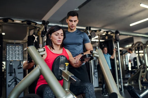 En kvinnlig senior med unga tränare gör styrka träning träning i gym. — Stockfoto