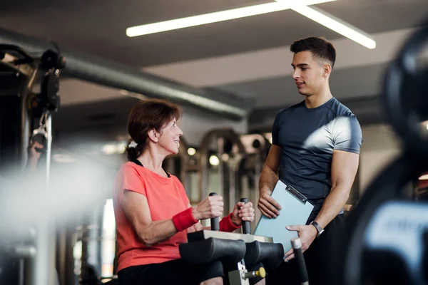 En kvinnlig senior med unga tränare gör styrka träning träning i gym. — Stockfoto