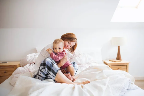 Une jeune mère avec une petite fille assise à l'intérieur sur le lit le matin, jouant . — Photo