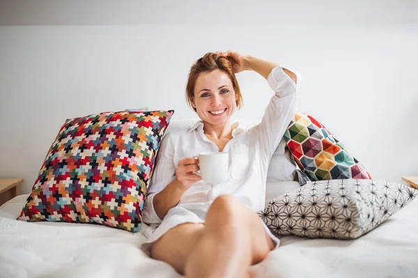 Young woman with night shirt sitting indoors on bed in the morning, holding cup. — Stock Photo, Image