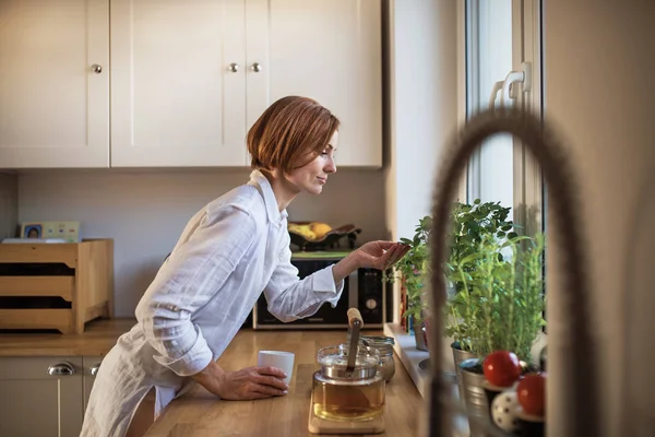 A young woman with cup of coffee standing indoors in kitchen, looking at plant. — Stock Photo, Image
