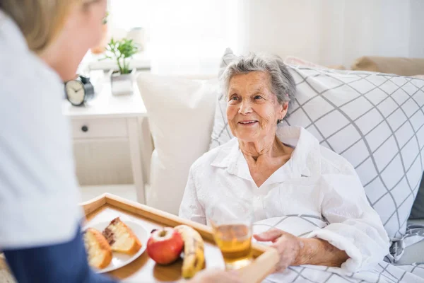 Un visitante de la salud llevando el desayuno a una anciana enferma acostada en la cama en casa . — Foto de Stock