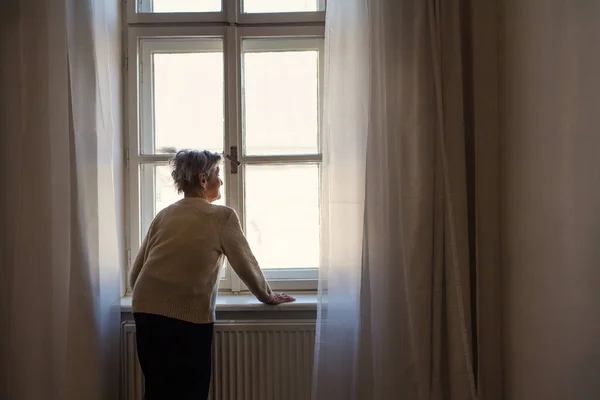Un ritratto di una donna anziana in piedi a casa, che guarda fuori da una finestra . — Foto Stock