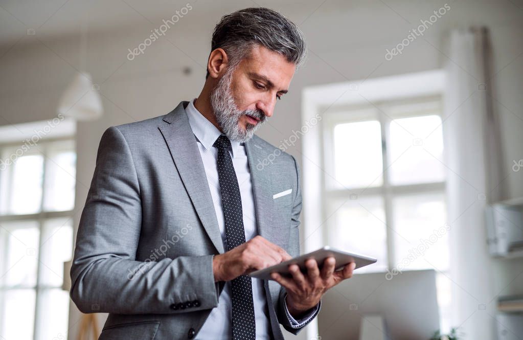 A mature businessman standing in an office, using tablet.