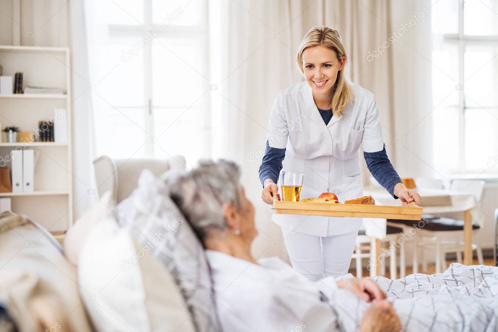 A health visitor bringing breakfast to a sick senior woman lying in bed at home.