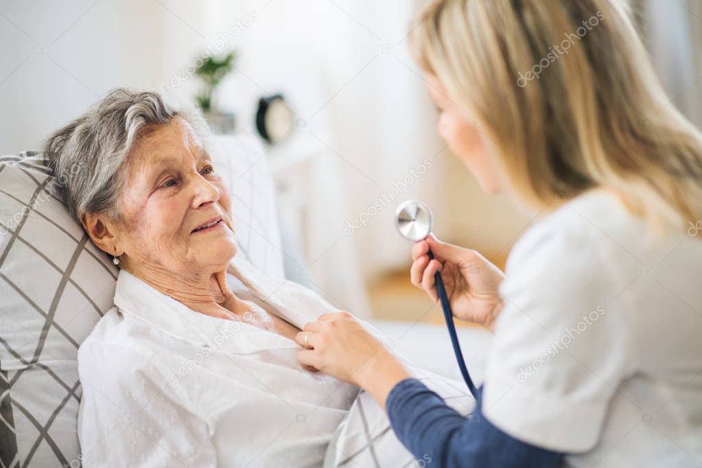 A health visitor examining a sick senior woman lying in bed at home with stethoscope.