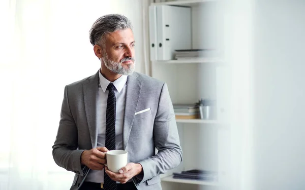Un uomo d'affari maturo in piedi in un ufficio, con in mano una tazza di caffè. Copia spazio . — Foto Stock