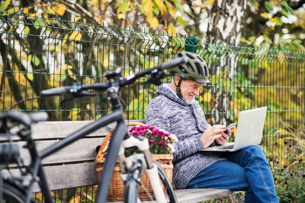 Hombre mayor con electrobicicleta sentado en un banco al aire libre en la ciudad, utilizando un ordenador portátil y un teléfono inteligente . — Foto de Stock