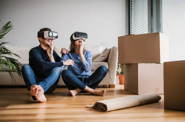 Una joven pareja con gafas VR sentada en el suelo, mudándose a un nuevo hogar . — Foto de Stock